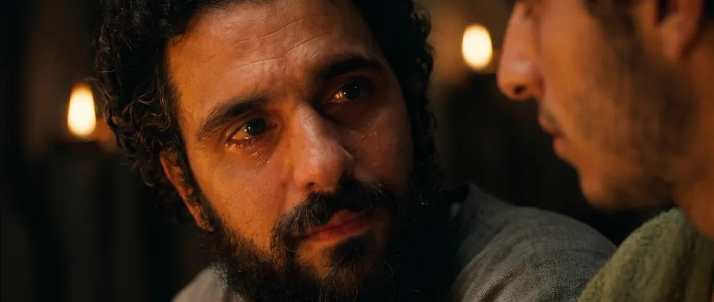 A bearded man in tears beside a younger man, by candlelight